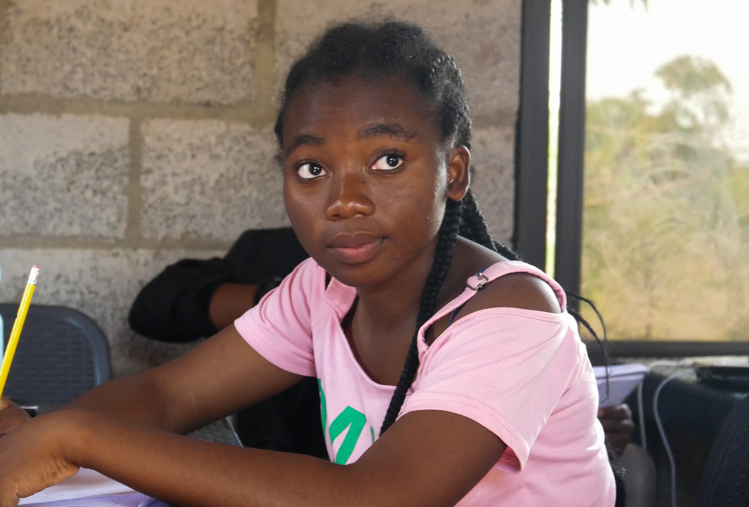 Mercy, a female student wearing a pink top sits at a desk with a notebook. She is looking just past the camera.